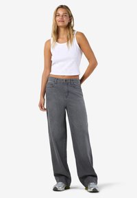 Hymyilevä nainen seisoo valkoisessa hihattomassa lyhyessä topissa, löysissä harmaissa farkuissa ja hopeisissa tennareissa valkoisella taustalla.