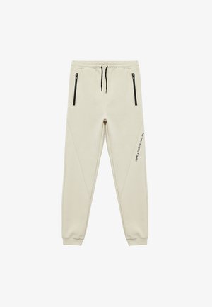 Beige joggers van zachte stof, met zwarte ritszakken en een taps toelopend ontwerp, voorzien van subtiele zijkanten en bedrukte details.