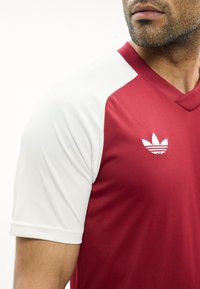 Mand iført en rød og hvid kortærmet skjorte med hvidt Adidas trefoil-logo på brystet, synlig fra hals til midt på armen.