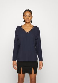 Top de manga longa azul-marinho com decote em V, apresentando um design texturizado e costuras subtis. Combinado com uma saia preta justa com dois rasgos.