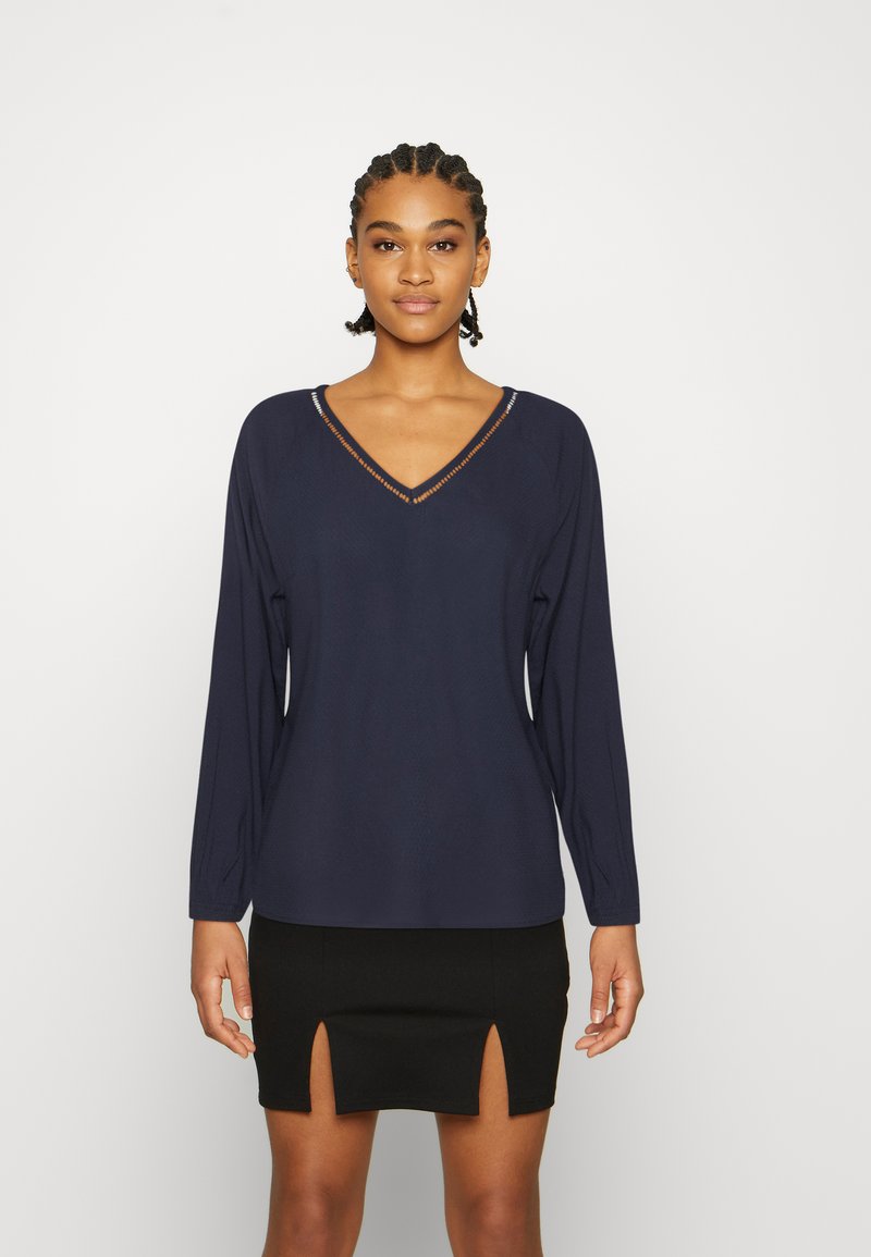 Top de manga longa azul-marinho com decote em V, apresentando um design texturizado e costuras subtis. Combinado com uma saia preta justa com dois rasgos.