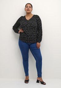 Blouse noire à manches longues avec motif floral violet, décolleté en V ; associée à un jean skinny bleu et des talons blocs noirs ouverts.