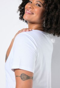 Biała bawełniana koszulka z krótkimi rękawami, o okrągłym dekolcie i tatuażem na górnej części ramienia. Gładka tekstura na jasnym tle.