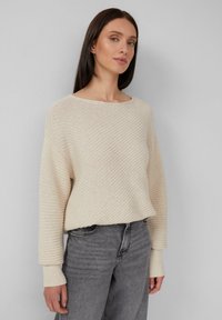 Donna che indossa un maglione a maglia testurizzato crema con maniche lunghe abbinato a jeans in denim grigi a vita alta su sfondo grigio.