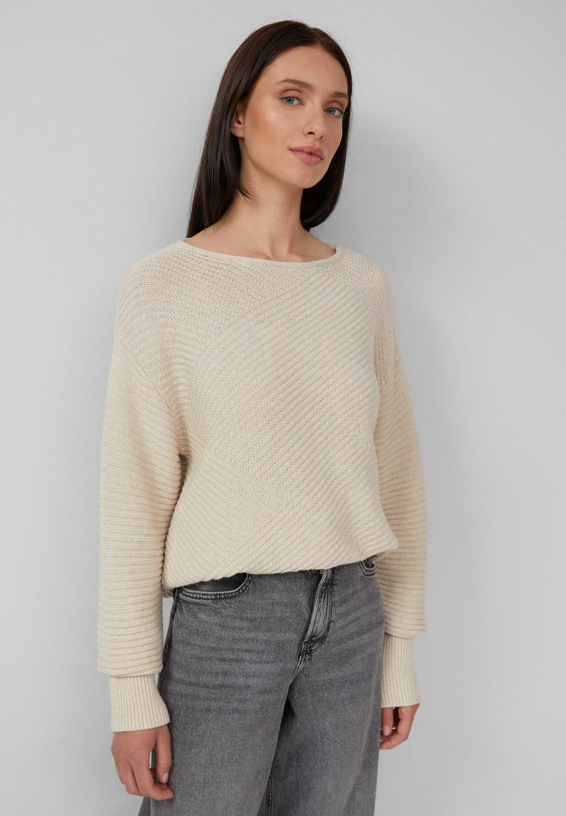 Donna che indossa un maglione a maglia testurizzato crema con maniche lunghe abbinato a jeans in denim grigi a vita alta su sfondo grigio.