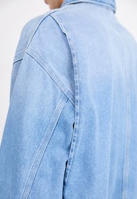 Hele sinine denim jakk, lõdva lõikega, nähtava õmblusega ja allapoole keeratud kraega. Tekstuur näeb välja pehme ja veidi kulunud.