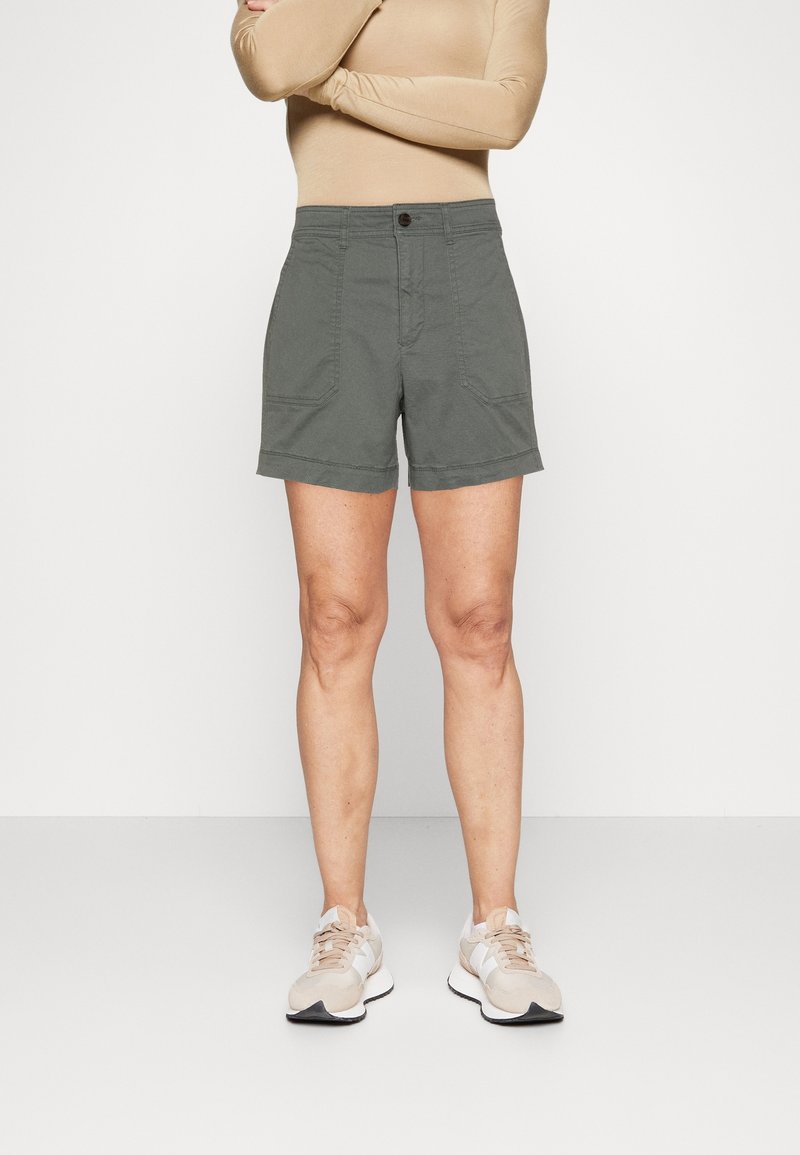 Pantalones cortos caqui hechos de una tela con una textura suave, con dos bolsillos delanteros y un dobladillo enrollado. Llevados con zapatillas deportivas beige.