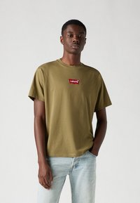 Olive green cotton t-shirt with a round neck and short sleeves, featuring a red Levi's logo patch on the chest. Worn with light denim jeans.