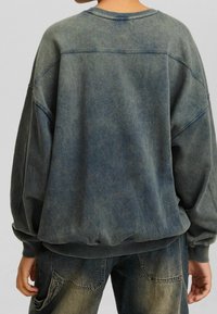 Sudadera de gran tamaño en azul grisáceo con cuello redondo, puños y dobladillo acanalados, que presenta una textura desgastada y detalles de costura visibles.