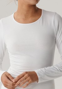 Zbliżenie osoby noszącej gładką białą koszulę z długim rękawem, dłonie delikatnie splecione na wysokości talii, zwrócona lekko w prawo.