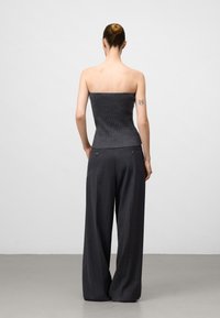 Femme avec les cheveux en chignon, portant un haut gris foncé sans manches et un pantalon large noir, debout avec les mains dans les poches, de dos, tatouage sur le bras gauche.