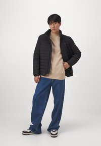 Giubbotto imbottito nero con cuciture a rombi e colletto rialzato, indossato sopra una felpa beige. Abbinato a jeans in denim blu a gamba larga e sneakers bianco/nero.