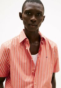 Camisa de botón de manga corta con rayas verticales rojas y azules, hecha de una tela ligera, con cuello clásico y detalle de bolsillo frontal.