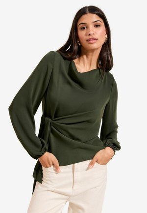 Frau mit langen dunklen Haaren, die goldene Ohrringe und ein olivgrünes Langarmshirt mit seitlicher Bindung trägt, kombiniert mit hellen Hosen.