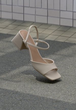 Sandalia beige de punta abierta con tacón ancho y tira delgada en el tobillo, inclinada hacia arriba sobre asfalto con fondo de pared de azulejos.