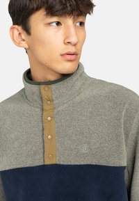 Fleece mikina v světle šedé barvě s tmavě modrou dolní částí, s vysokým límcem, knoflíkovou légou a logem na hrudi.