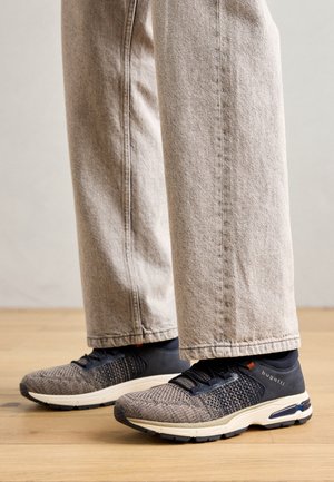 Sportliche Schuhe mit einem strukturierten grauen und schwarzen Strickobermaterial, schwarzem Fersenbereich und einer weißen Sohle mit blauen Akzenten, getragen mit hellbeigen Jeans.