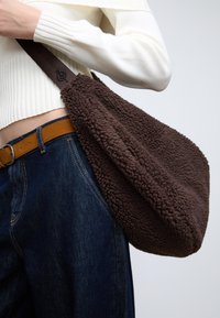 Hnedá ramenná taška zo shearlingu s mäkkou textúrou, vybavená pevnou hnedou remienkom a zaobleným tvarom, nosená spolu s tmavými džínsami.