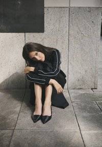 Abbigliamento nero con strisce bianche, texture liscia. Il modello è seduto su una superficie di pietra, indossando scarpette nere a punta, capelli sciolti, braccia attorno alle ginocchia.