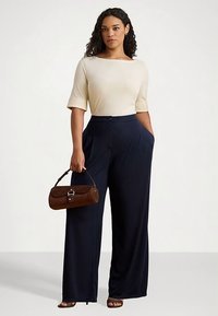 Bēša īsroku krekls kopā ar tumši ziliem plaša piegriezuma biksēm ar sānu kabatām un pogas aizdari. Tur rokās brūnu rokassomu.