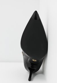 Chaussure noire à talon haut avec un orteil pointu, texture en cuir lisse, talon aiguille fin et un accent doré sur le côté.