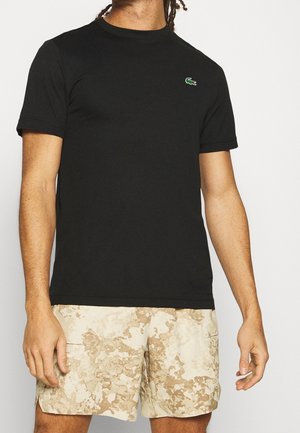 Homme portant un t-shirt noir à manches courtes avec un petit logo vert et un short beige à motif camouflage sur un fond uni.