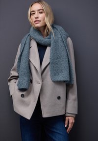 Lysegrå dobbeltknappet uldfrakke med sorte knapper, kombineret med et tekstureret turkis tørklæde. Mørkeblå top og mørke jeans fuldender outfittet.