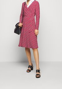 Czerwona sukienka w stylu kopertowym z długimi rękawami, białym kołnierzykiem i geometrycznym wzorem. Noszona z czarnymi sandałami na słupku. Czarna torebka z frędzlami.