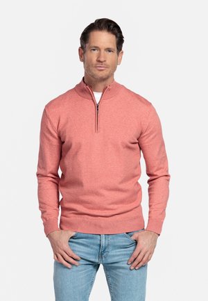 Man staat met handen in zakken, draagt een koraalroze zip-up trui en lichtblauwe spijkerbroek tegen een effen witte achtergrond.