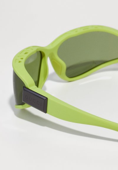 Gafas de sol con un marco de goma mate verde y lentes oscuros. Presentan una placa rectangular negra con el logo en la patilla y un diseño deportivo.