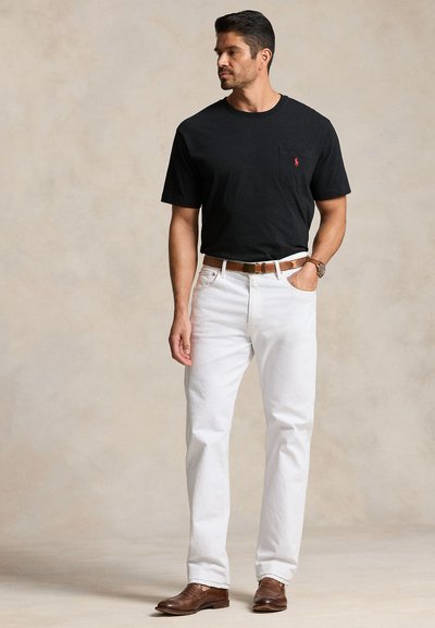 Moški, ki nosi črno majico s kratkimi rokavi, bele hlače, rjavi pas in rjave čevlje, stoji z eno roko v žepu in gleda vstran.