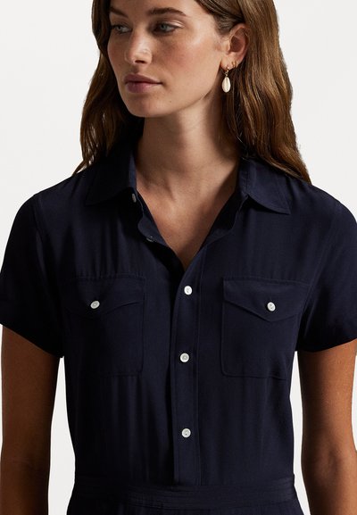 Vrouw met lang bruin haar in een marineblauw kortemouw overhemd met twee borstzakken en schelpenoorbellen.