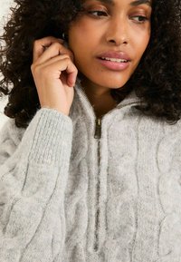 Hellgrauer, gestrickter Pullover mit einem Zopf-Muster, ausgestattet mit einem Reißverschlusskragen und gerippten Bündchen. Weiche Textur mit einem fusseligen Erscheinungsbild.