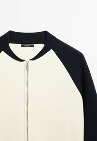 Pull zippé noir et blanc avec col et manches côtelés noirs, corps blanc et fermeture éclair argentée au centre devant.