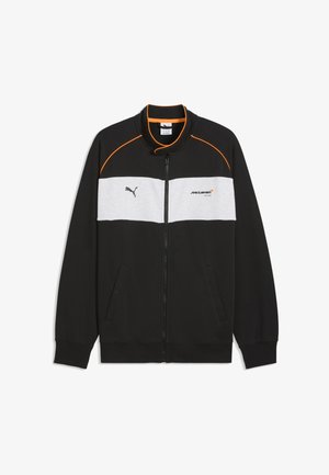 Czarna kurtka na zamek błyskawiczny z białym panelem na piersi, pomarańczowymi lamówkami na ramionach, logo Puma po lewej stronie i logo McLaren Racing po prawej stronie klatki piersiowej.
