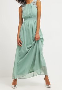 Maximekko vaaleassa mintunvihreässä, jossa on laskostettu bodice, pitsikoristeet olkapäillä ja virtaava kangas, jossa on kevyt kiilto. Hopeiset korot ovat näkyvissä.