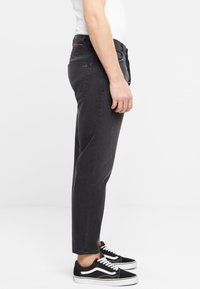 Pantalones de mezclilla negros con corte recto, desgaste sutil y bolsillos frontales. Combinados con zapatillas negras y blancas.