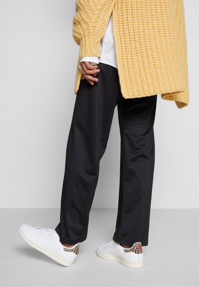 Lockere schwarze Hose, gestrickte gelbe Strickjacke und weiße Sneakers mit einem Leopardenmuster an der Ferse. Strickjacke mit Struktur, lässiger Look.