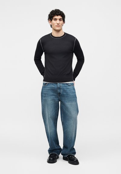 Jeune homme aux cheveux bouclés, debout, les mains derrière le dos, portant une chemise noire à manches longues, un jean bleu large et des chaussures noires.