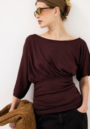 Donna con capelli biondi raccolti in uno chignon attorcigliato, che indossa occhiali da sole color ambra e un top scuro color bordeaux arricciato, che tiene in mano una grande borsa intrecciata beige.