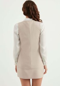 Beige ärmlös klänning med en slät textur, som har en figursydd rygg och en vit långärmad skjorta under med knapppropp.