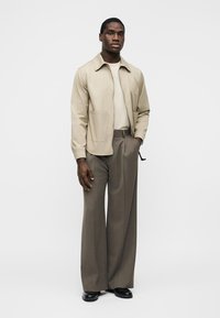 Άνδρας που στέκεται φορώντας μπεζ μπουφάν, ανοιχτό μπεζ πουκάμισο, καφέ παντελόνι με φαρδιά μπατζάκια και μαύρα παπούτσια, πάνω σε μονόχρωμο λευκό φόντο.