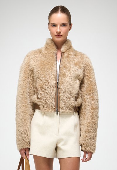 Béžová nadýchaná umelá kožušinová bomber bunda so zipsom vpredu a dvoma bočn�ými vreckami, skombinovaná so svetlými strihovými šortkami. Hladká, textúrovaná látka.