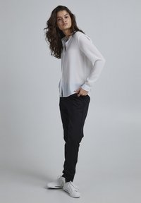 White long-sleeve blouse with a button-up front, paired with black tapered pants and white sneakers. Smooth fabric, relaxed fit, minimal design.