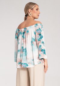 Bluzka typu off-the-shoulder z lekkiego, przezroczystego materiału w kwiatowy wzór w odcieniach turkusu i delikatnego różu. Rękawy o długości trzy czwarte, luźny krój.
