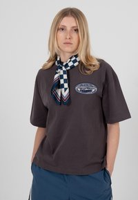 Jeune personne aux cheveux blonds longs portant un t-shirt sombre ample, une écharpe à carreaux bleue et un pantalon bleu, les mains dans les poches.