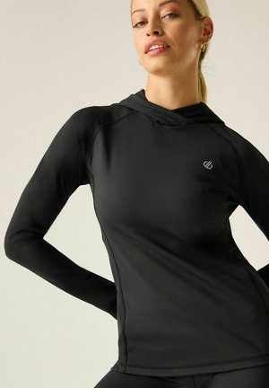 Young woman wearing a fitted black long-sleeve hoodie with small logo on chest, posing with hands on hips against a plain background.