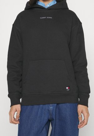 Osoba nosząca czarną bluzę z kapturem Tommy Jeans z przednią kieszenią na niebieskiej koszuli z kołnierzykiem i niebieskich dżinsach, stojąca na jednolitym tle.
