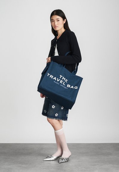 Saco de lona azul-marinho com grande texto branco "THE TRAVEL BAG MARC JACOBS." Apresenta duas alças robustas e uma forma estruturada.