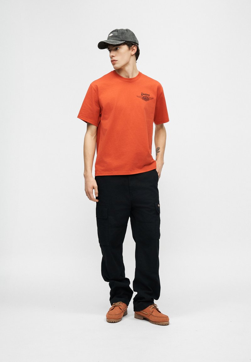 Junger Mann trägt ein orangefarbenes Dickies-T-Shirt, schwarze Cargo-Hose, graue Kappe und braune Schuhe, steht mit einer Hand in der Tasche und schaut nach links.
